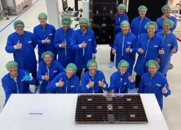 A group of 16 people in blue lab coats and green hairnets stand in a laboratory, giving thumbs-up gestures and smiling. In front of them are two solar panel arrays labeled 