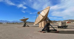 Two large satellite dishes are set up in a desert landscape under a blue sky with scattered clouds. Each dish is mounted on a sturdy base, and next to one of the dishes, a control unit bears the marking: 