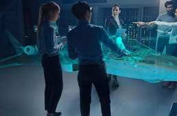 A group of five people in a futuristic control room observe and interact with a large, blue holographic model of an advanced aircraft, part of the Future Combat Air System (FCAS). The background features illuminated computer panels and screens that seamlessly integrate with the Internet of Military Things.